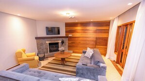 A modern living room with a stone fireplace, a sofa set, a coffee table, and a television mounted on the wall. - Villa Hegus (Campos do Jordão)