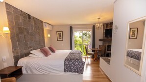 A bedroom with a bed, a stone wall, a balcony with a table and chairs, and a fireplace. - Villa Hegus (Campos do Jordão)