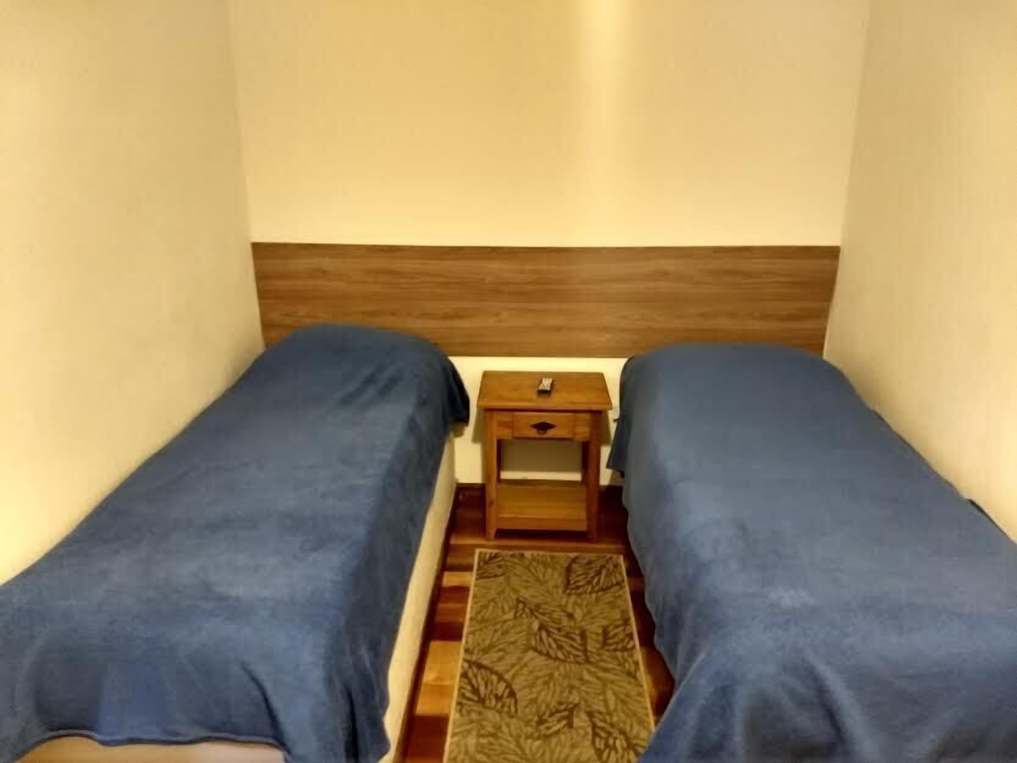 A small, single-room hotel room with two single beds, a wooden nightstand, and a patterned rug.