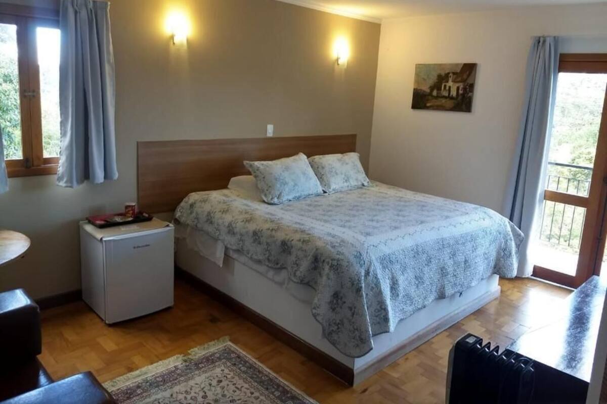 Un dormitori amb un llit, taules de nit, petita nevera, finestra amb cortines i un quadre a la paret.