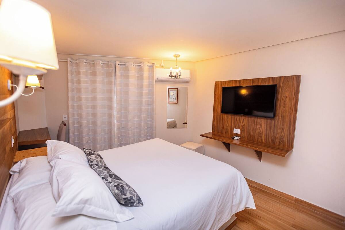 Habitació d'hotel amb un llit, televisió penjada, taula de nit i cortina de bany.