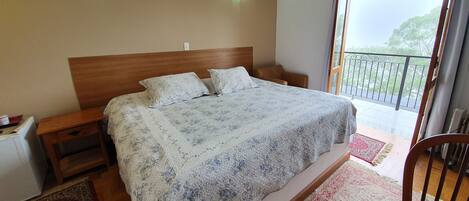 Un dormitorio con una cama grande, cabecera de madera, dos mesitas de noche, un perchero y un balcón con barandilla y vistas al verdor.