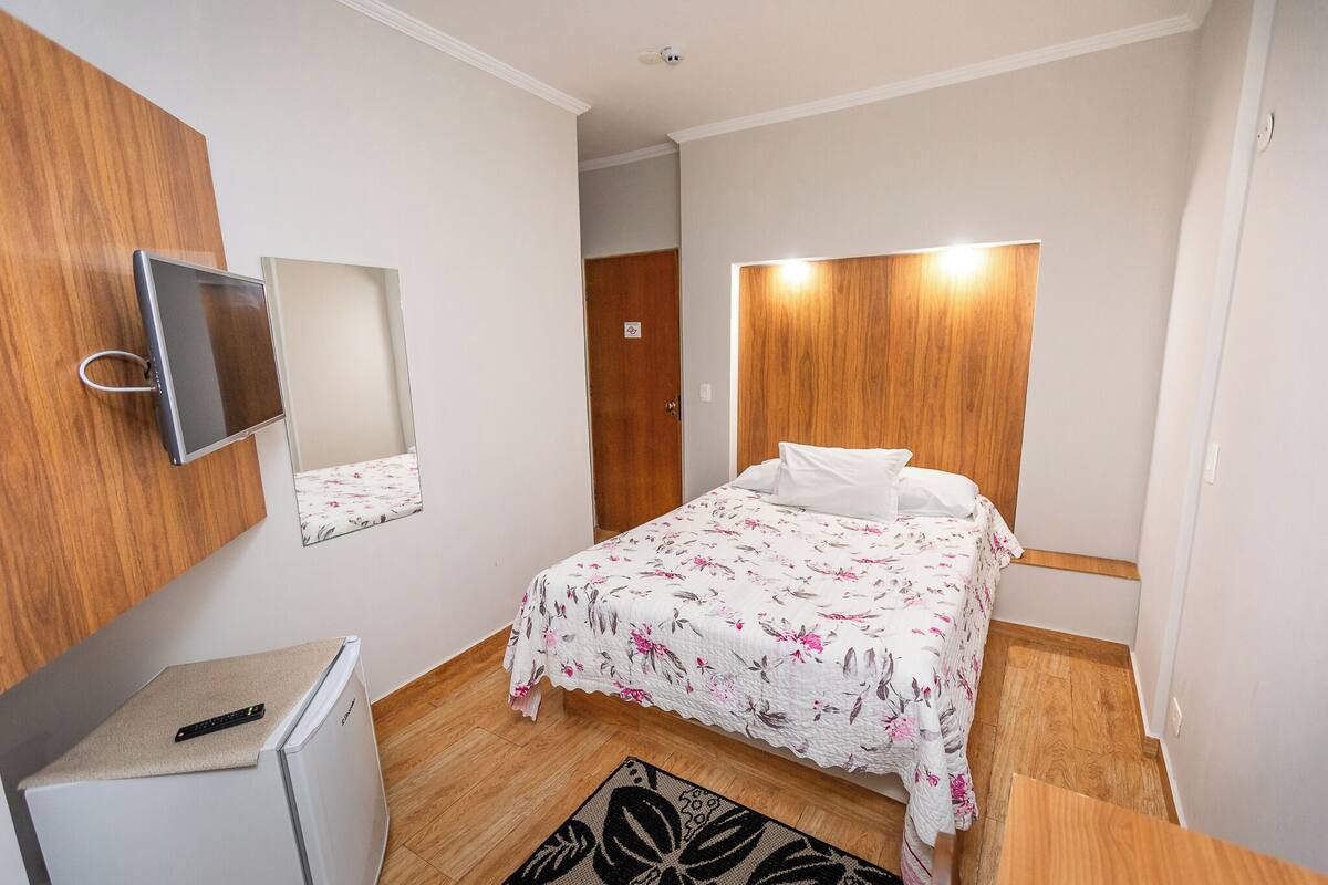 Un dormitori amb un llit, un petit frigorífic, un mirall i un armari de fusta.