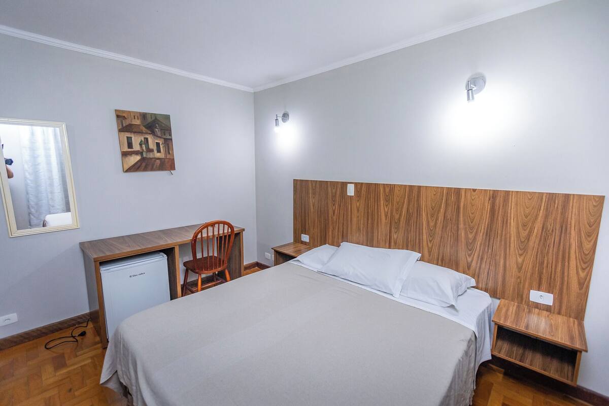 Habitació amb llit, escriptori, cadira, nevera petita i lluminària penjada a la paret.
