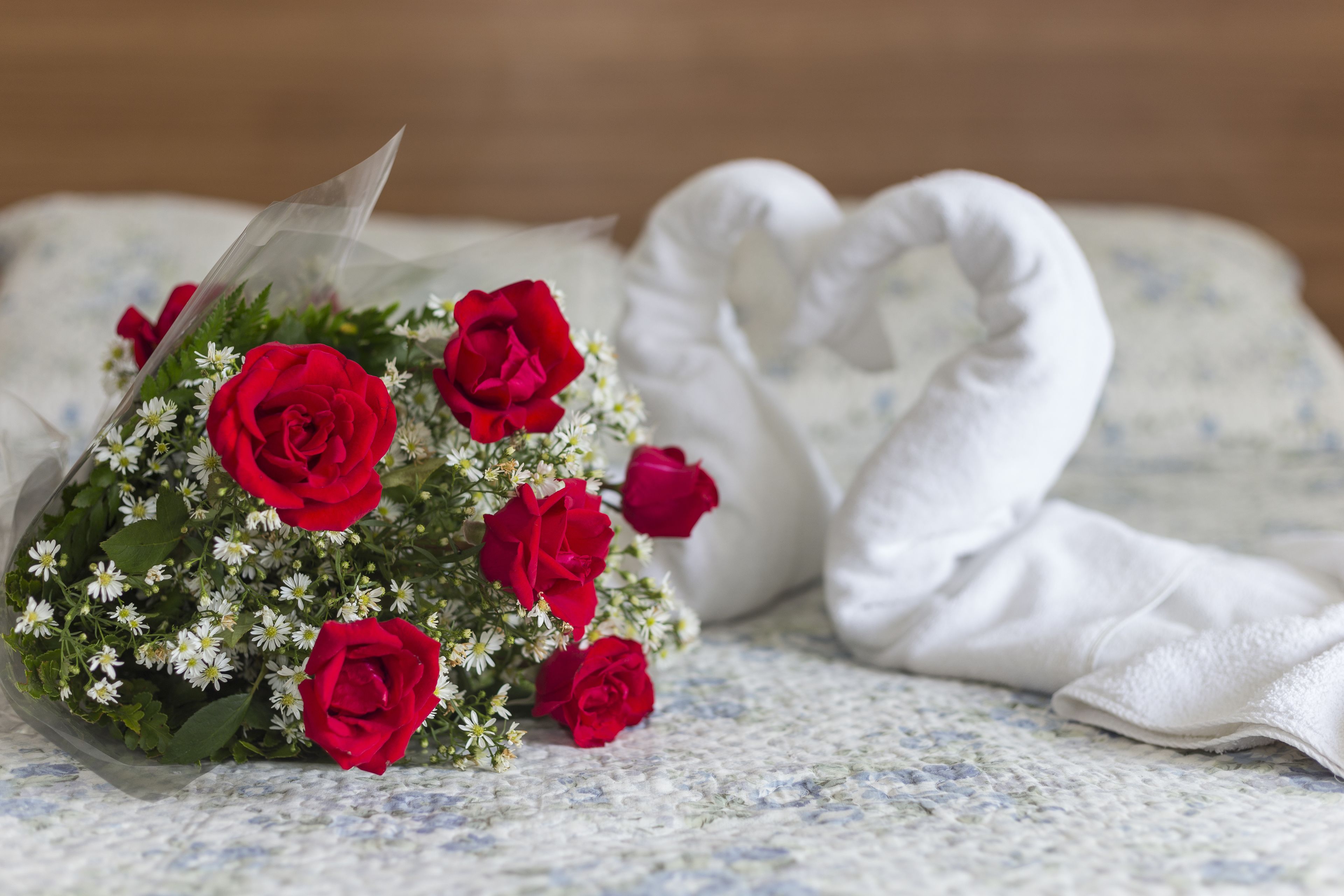 A bouquet of red roses and a white heart-shaped pillow on a bed with a floral-patterned bedspread.