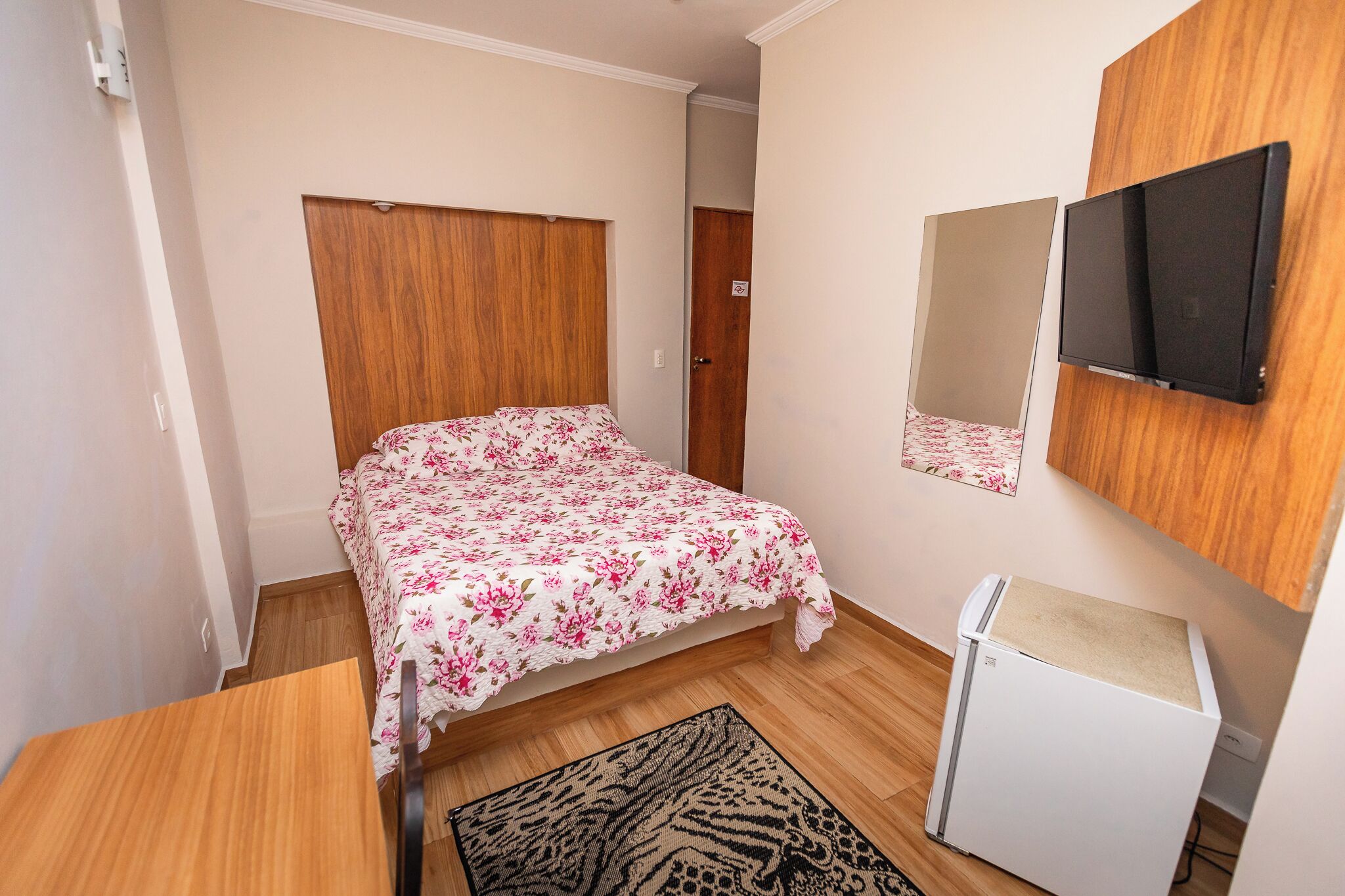 A small, neatly arranged bedroom with a bed, a wooden wardrobe, a small white cabinet, and a television mounted on the wall.