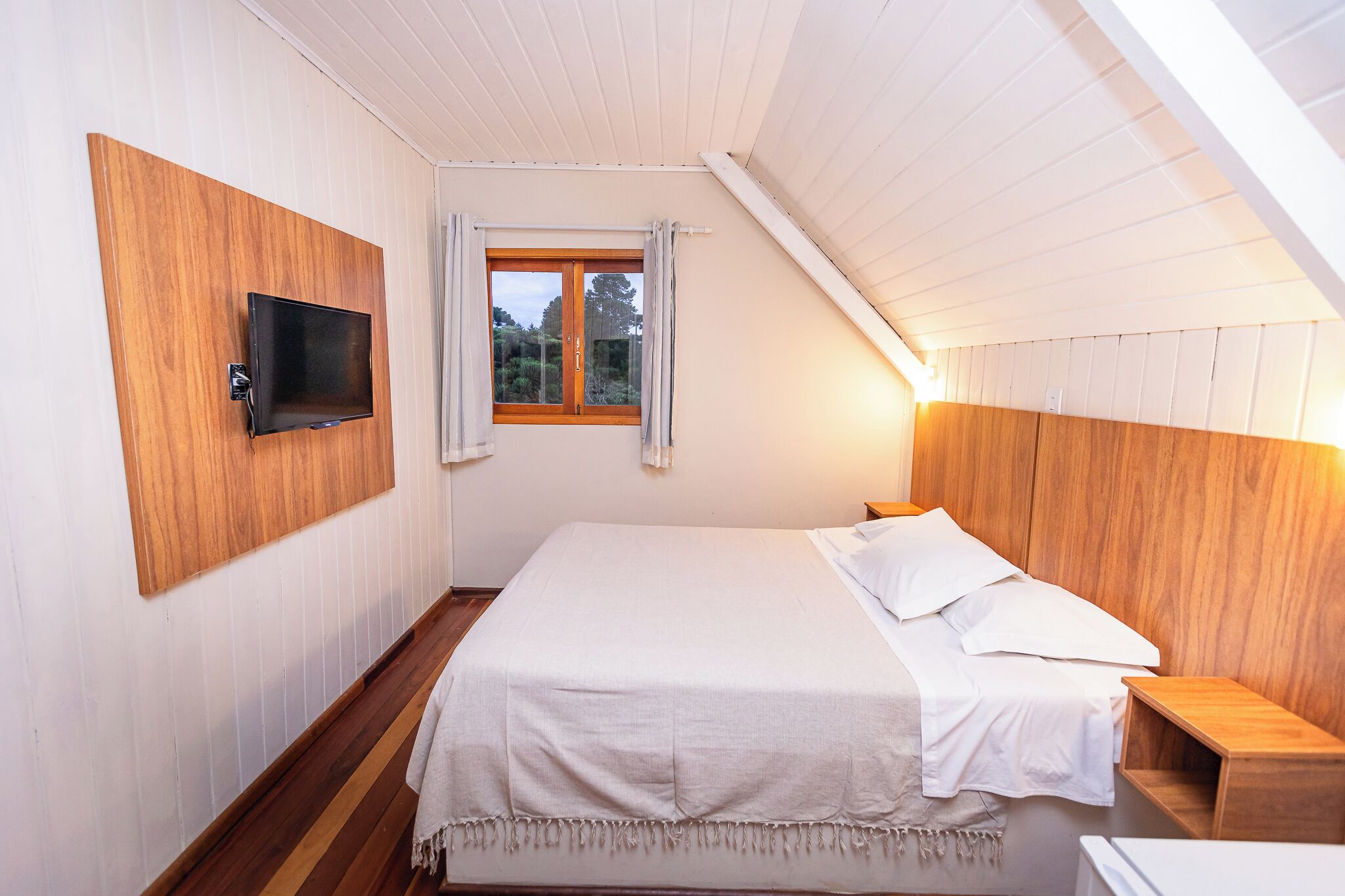 Un dormitorio con cabecera de madera, televisor montado en la pared, ventana con cortinas y mesita de noche.