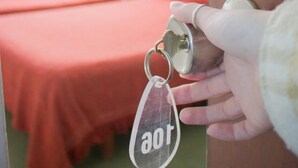 Una mano sostiene una llave con un llavero que dice 'aot' en una habitación de hotel con dos camas y un ventilador de techo.