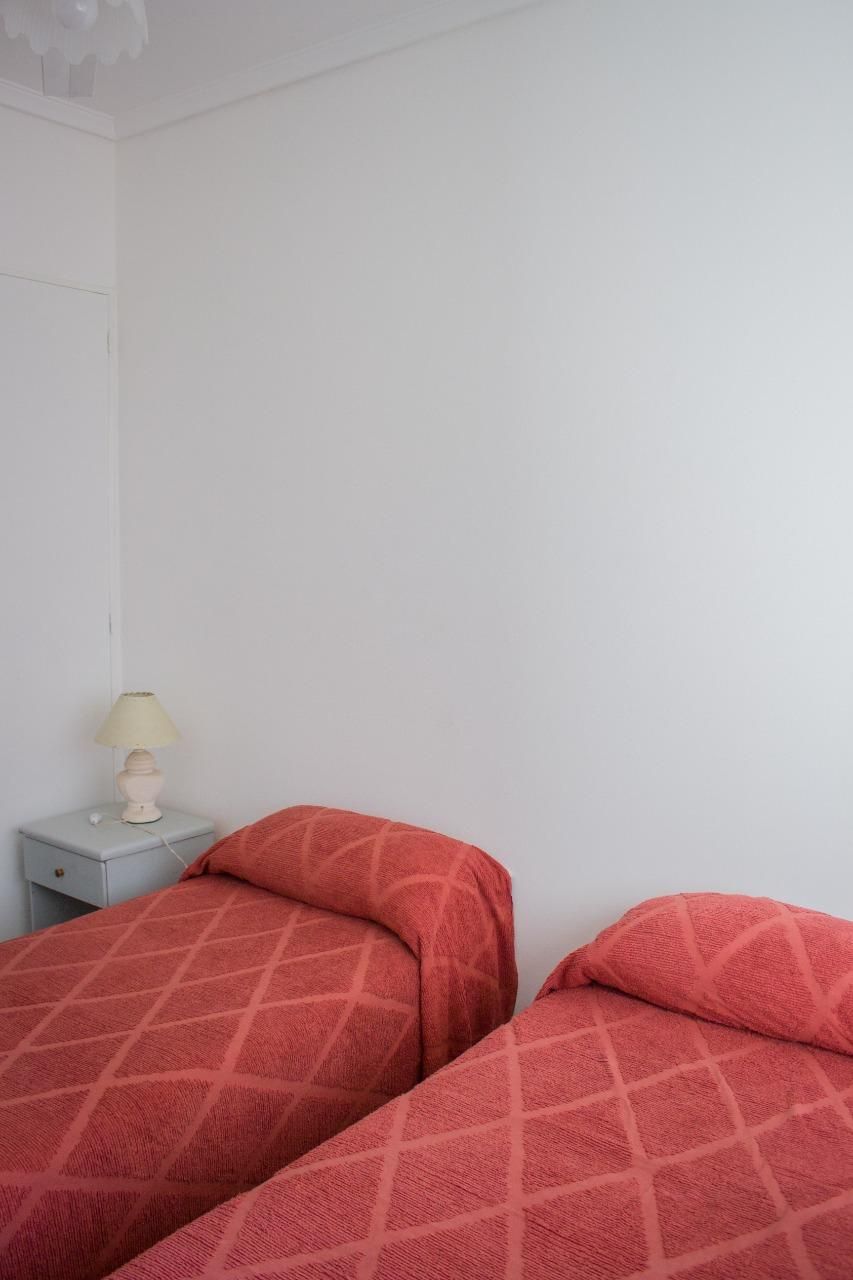 A room with two beds, a nightstand with a lamp, and a white wall.