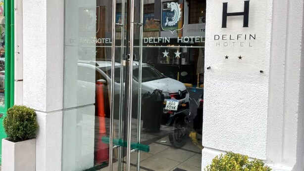 Exterior del Hotel Delfin, con entrada de vidrio, un coche estacionado afuera y dos macetas a cada lado de la entrada.