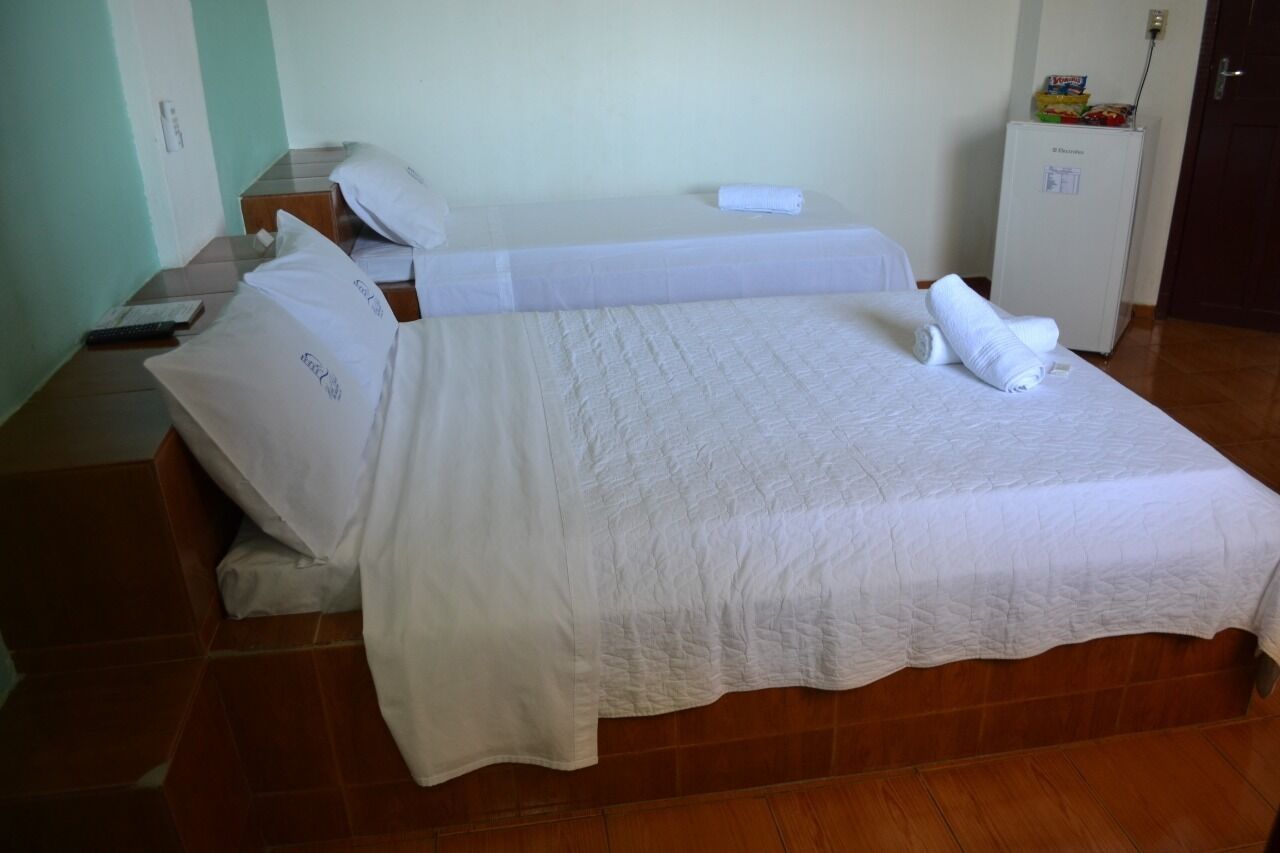 Una cama bien hecha con sábanas y almohadas blancas, una mesita de noche con un control remoto y un refrigerador en la esquina.