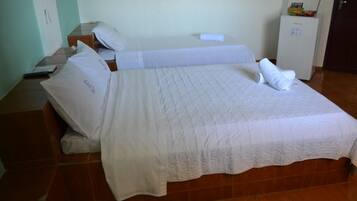 Una cama bien hecha con sábanas y almohadas blancas, una mesita de noche con un control remoto y un refrigerador en la esquina.