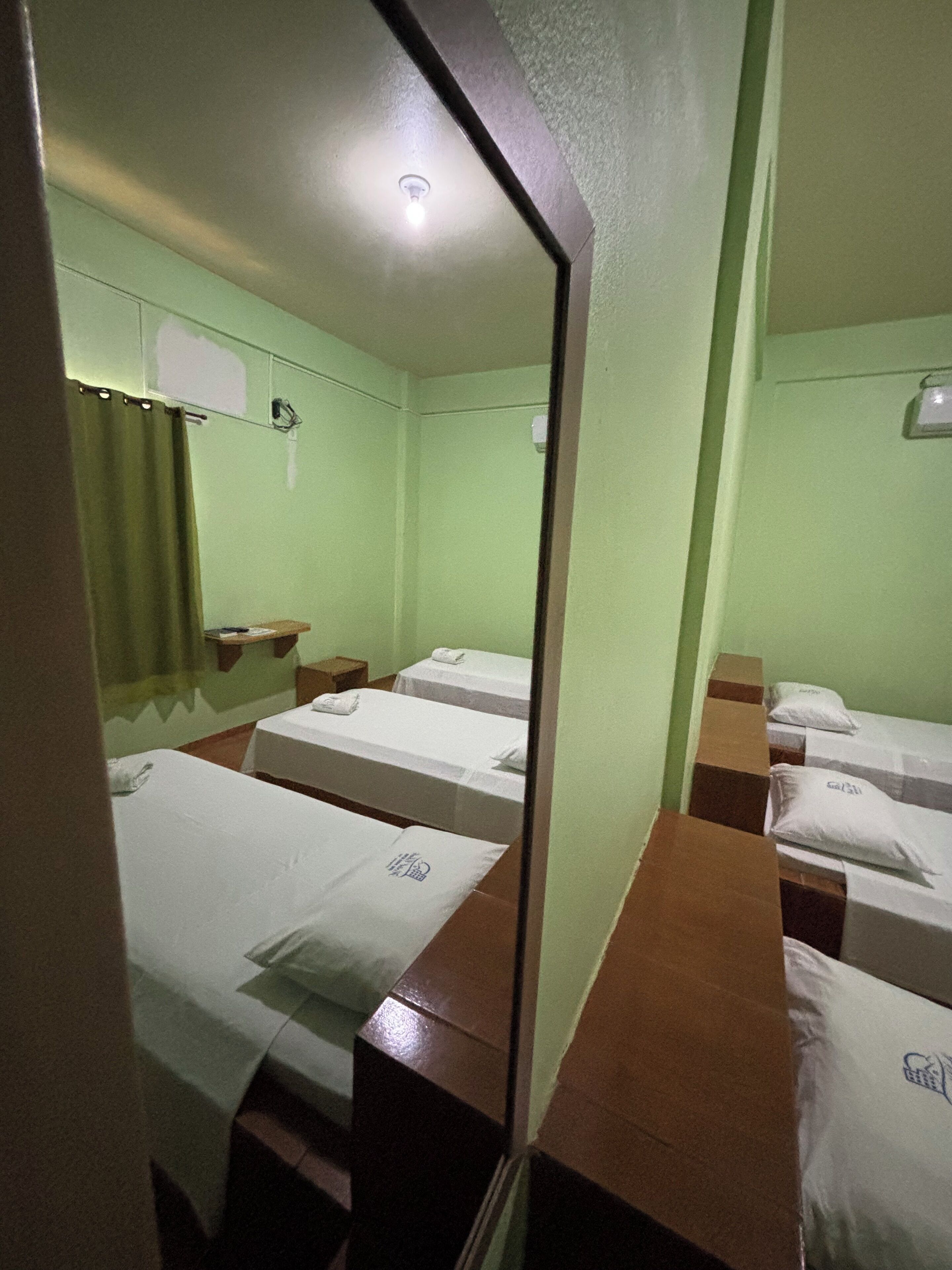 Habitación de hotel con tres camas individuales, una mesita pequeña, un espejo y una lámpara en el techo.