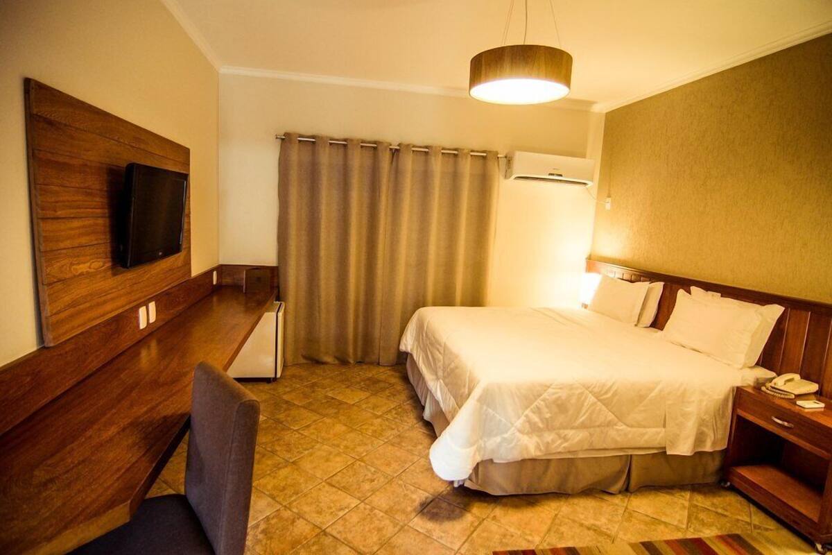 A hotel room with a bed, a desk, a chair, a television mounted on a wooden panel, a lamp, and a curtain.