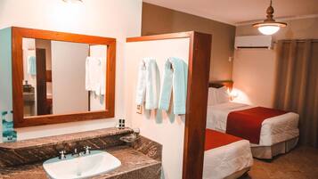 Quarto de hotel com lavatório de mármore, um espelho grande, duas camas com roupa de cama vermelha e branca, e um ventilador de teto.