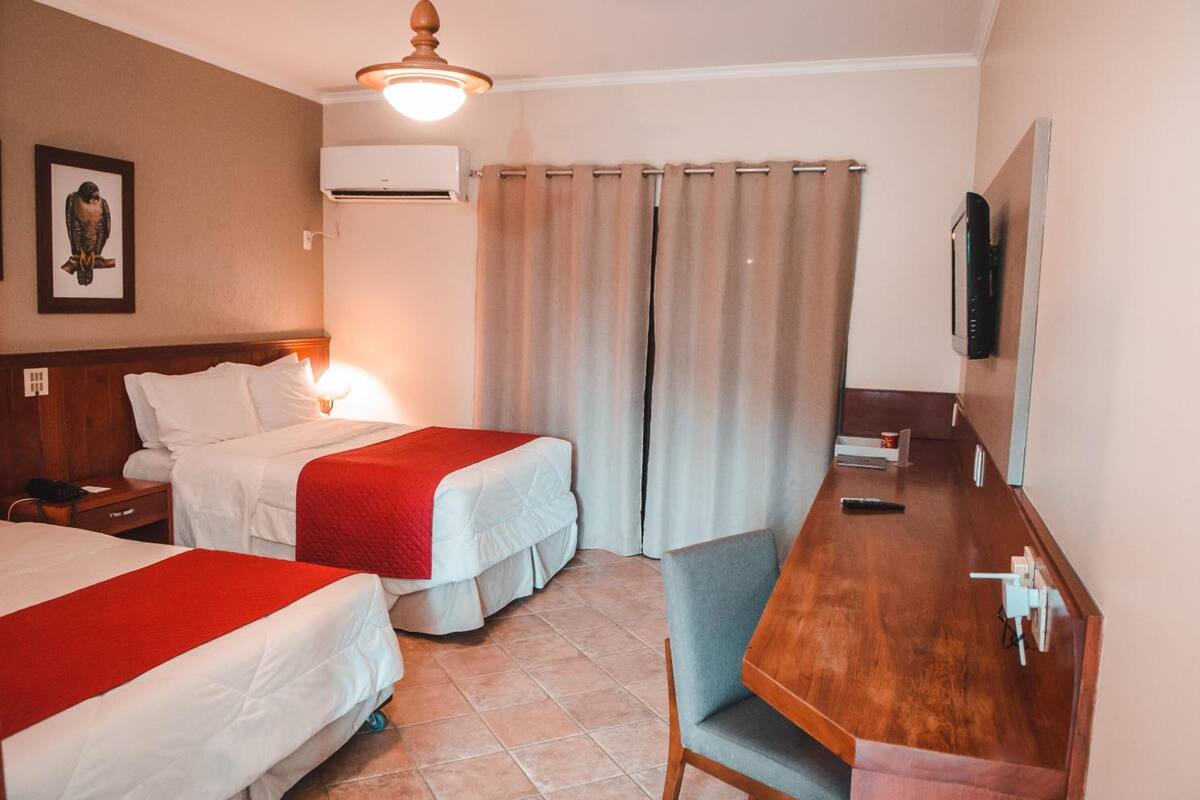 A hotel room with two beds, a wooden desk, a chair, a television, and a window with curtains.