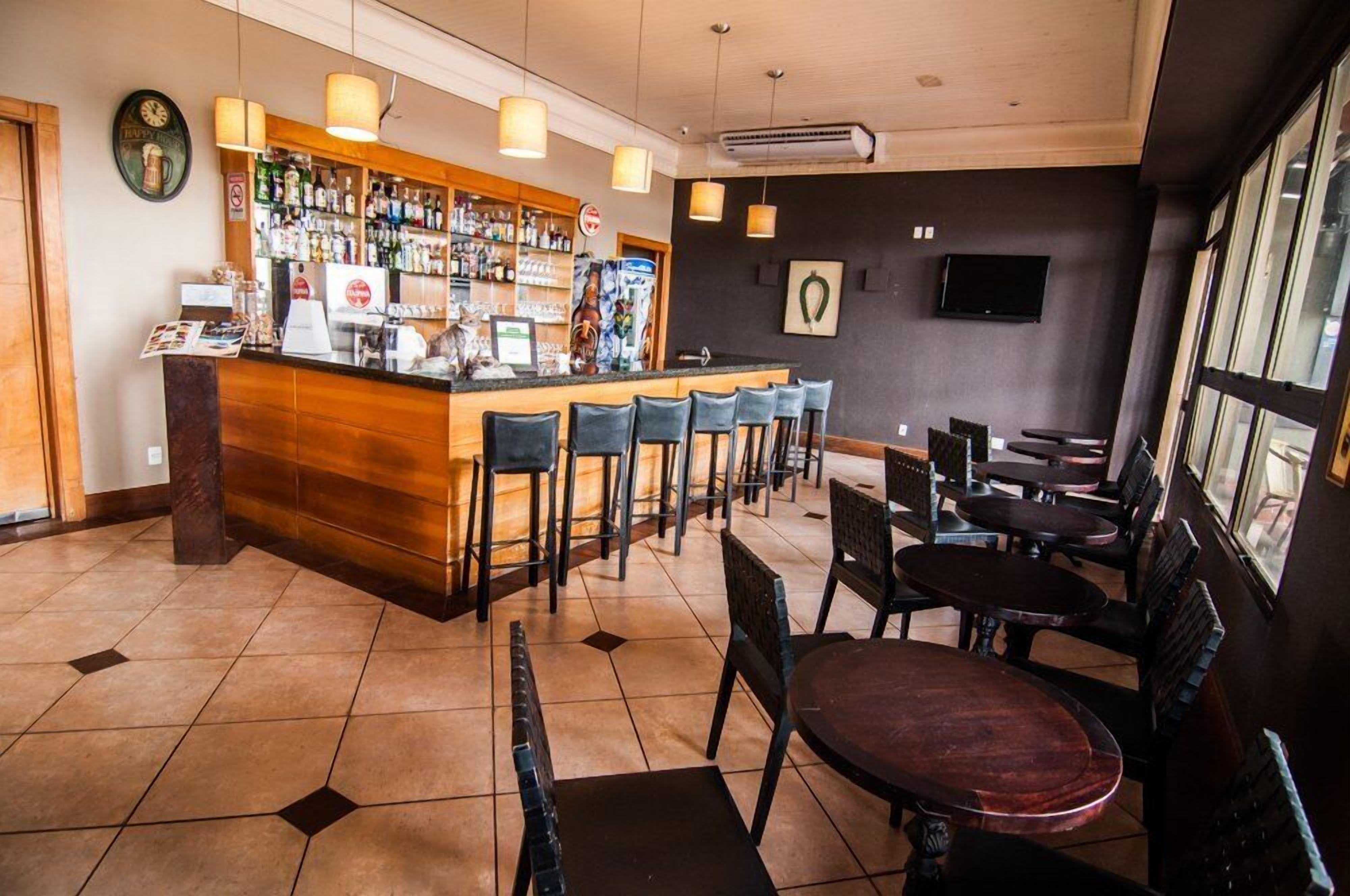 Zona de bar con mostrador de madera, varios taburetes, y una variedad de botellas. Hay mesas y sillas para sentarse, y un televisor montado en la pared.
