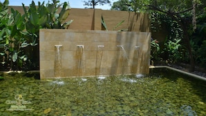 Un mur de pierres sur lequel de l’eau s’écoule en cascade pour se déverser dans un bassin limpide parsemé de galets.