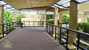 Une terrasse en bois dotée d’un toit en verre, entourée de verdure et agrémentée d’un élément aquatique.