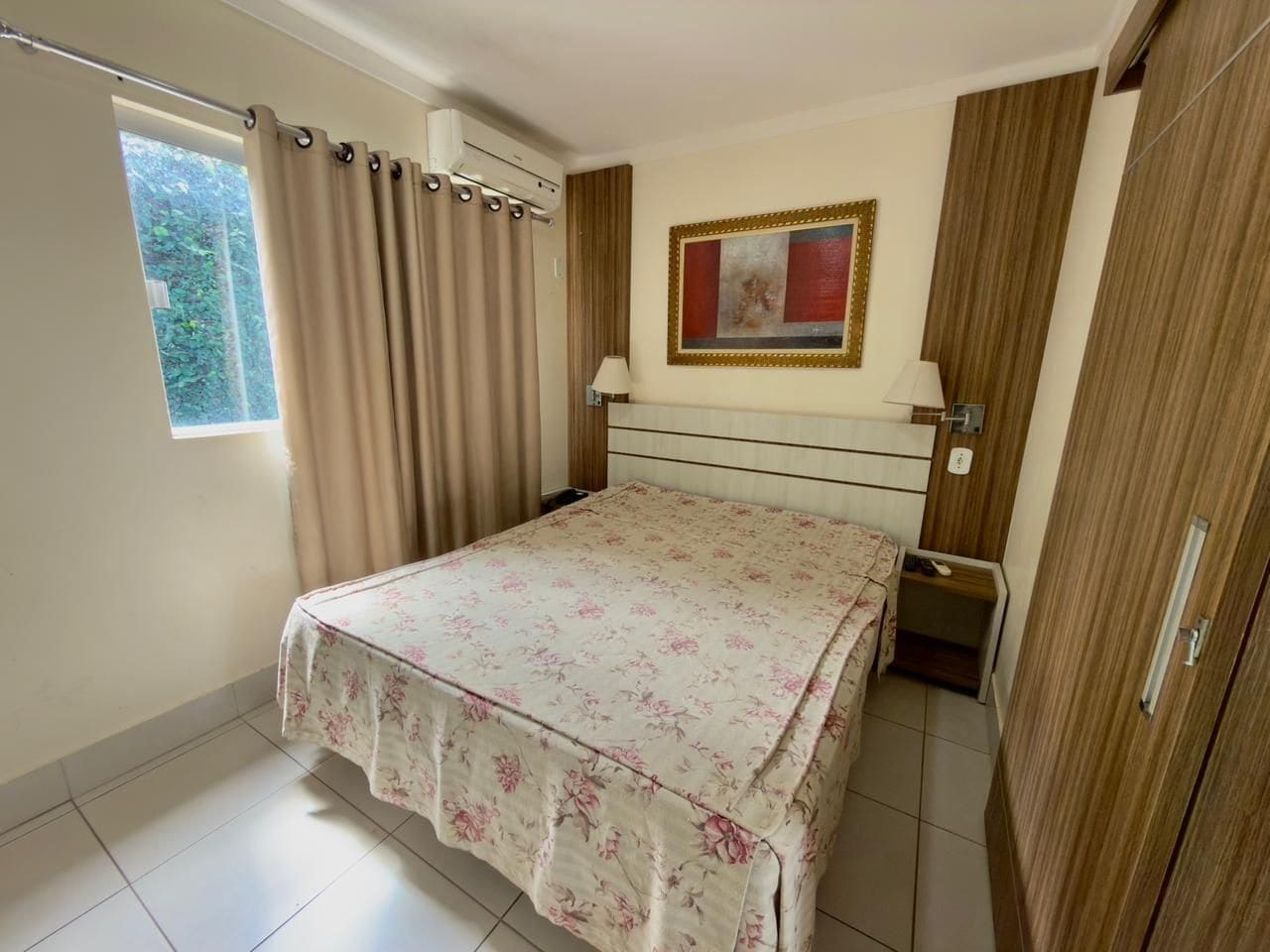 A bedroom with a bed, curtains, a nightstand, a lamp, a wardrobe, and a window with a view of greenery.