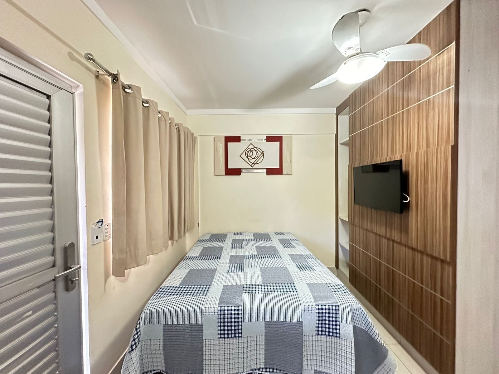 A bedroom with a bed, a ceiling fan, a TV mounted on the wall, and a door with a curtain.