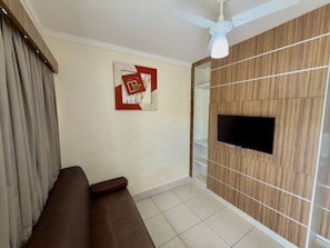A living room with a brown sofa, a mounted TV, a ceiling fan, and a wall-mounted shelf.