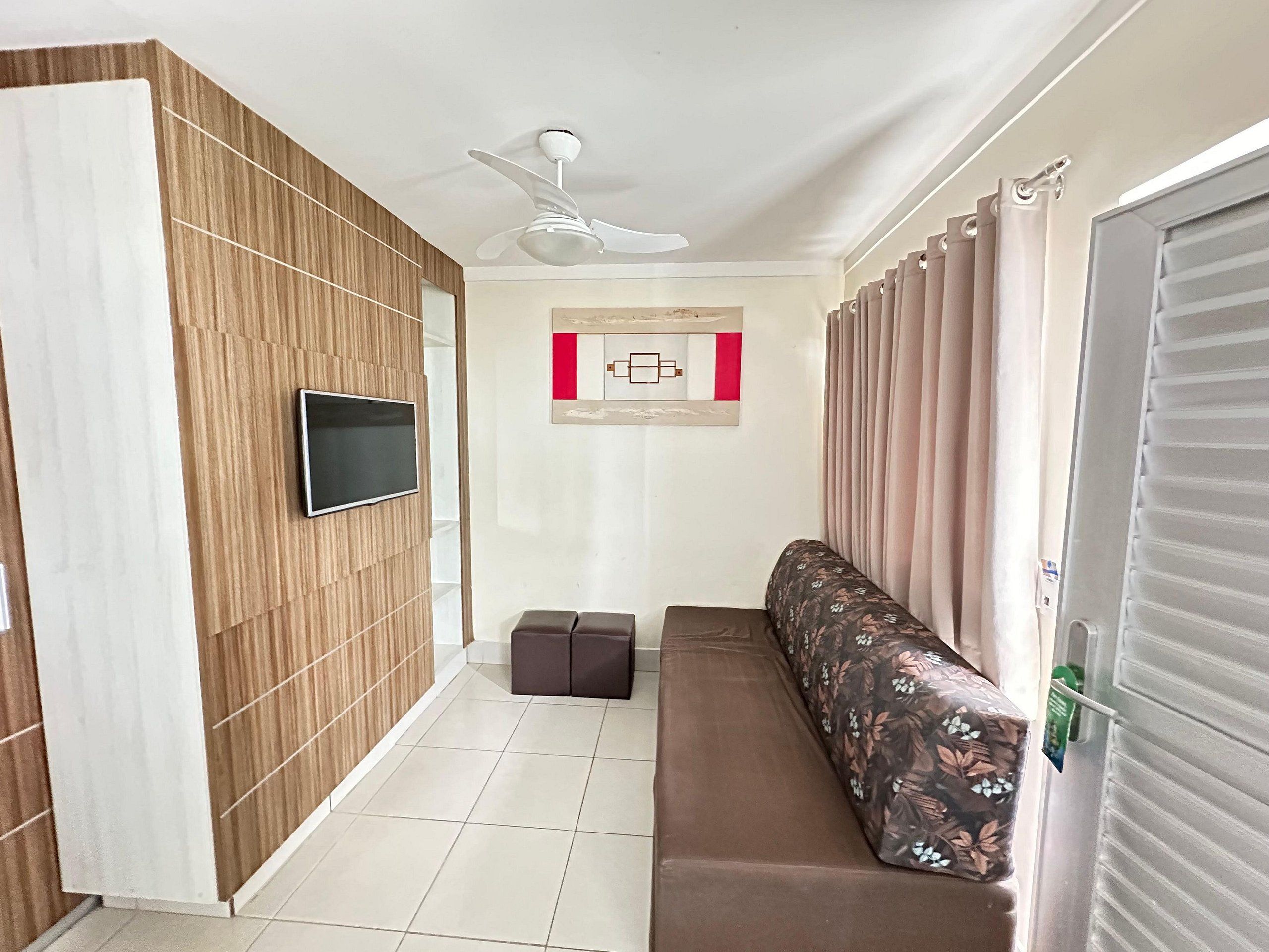 Un pasillo con ventilador de techo, televisor de pantalla plana montado en una pared de paneles de madera, un banco con tapicería floral y una puerta con ventana.