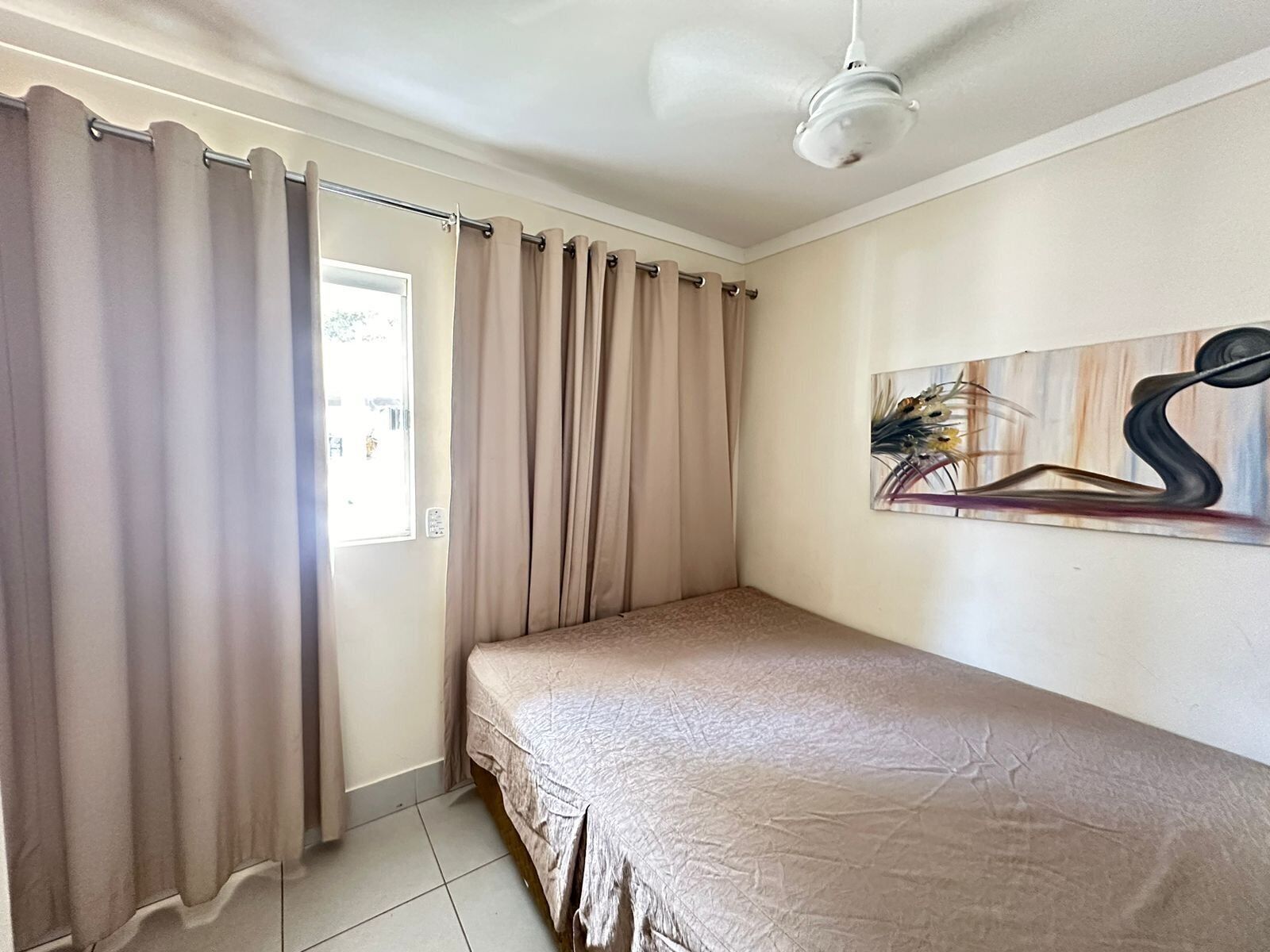 A bedroom with a single bed, beige curtains, a ceiling fan, and a painting on the wall.