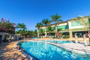 Una piscina con sillones de descanso y palmeras, rodeada por un edificio con grandes ventanales y un paseo cubierto.