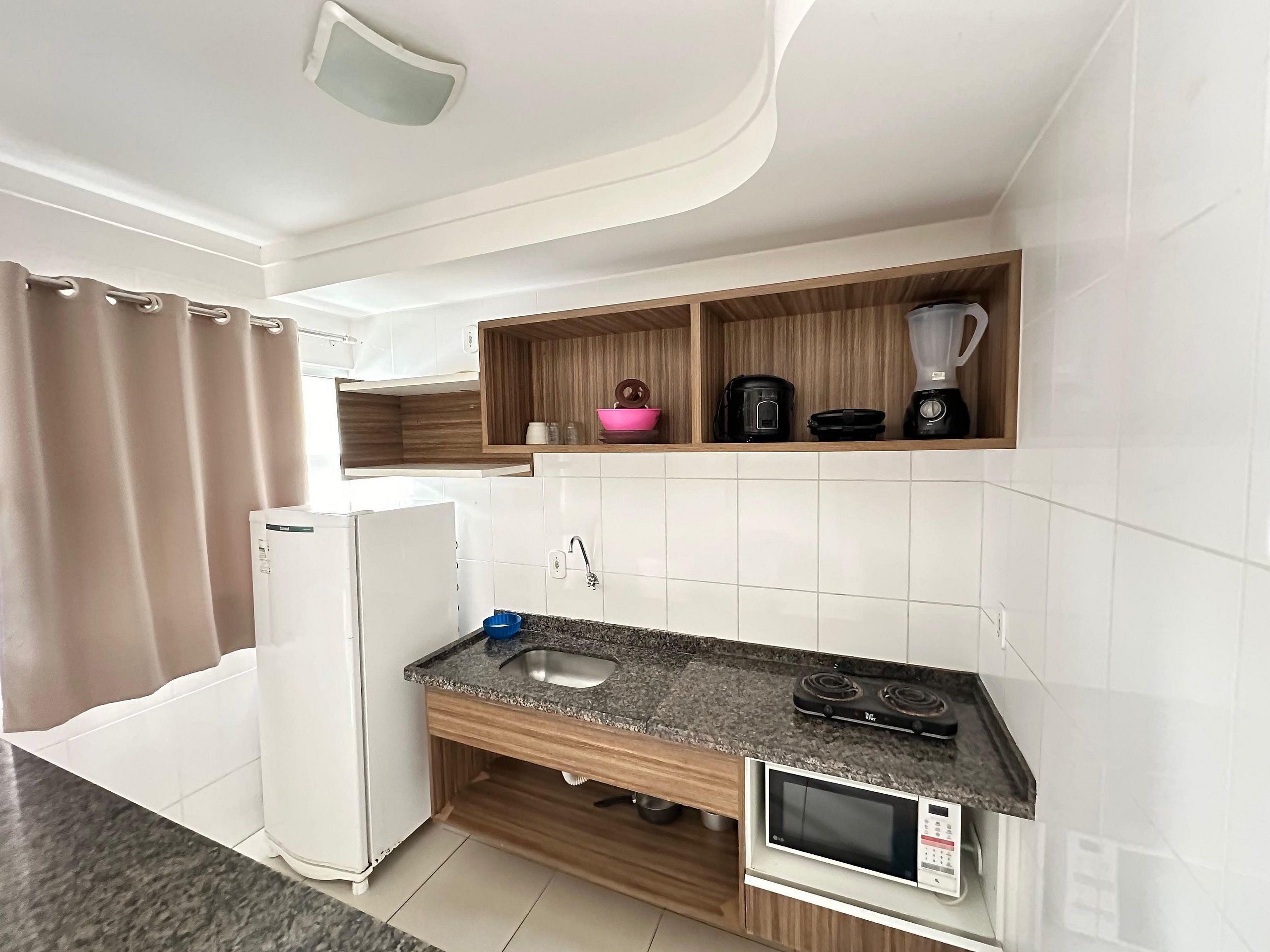 A compact kitchen with a built-in microwave, a sink, and a stove. There are wooden shelves above the counter holding a blender, a kettle, and a pink bowl.