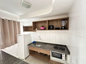 A compact kitchen with a built-in microwave, a sink, and a stove. There are wooden shelves above the counter holding a blender, a kettle, and a pink bowl. - Lacqua Di Roma I Via Caldas (Caldas Novas)