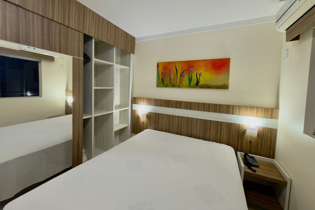A hotel room with a bed, a painting, a desk with a phone, and a wardrobe.