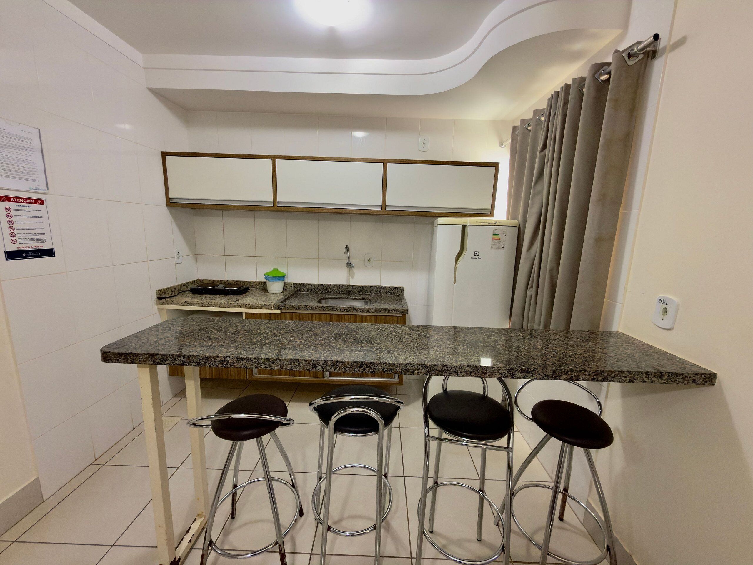 Una cocina con encimera de granito, un refrigerador, un lavamanos y tres taburetes de bar.