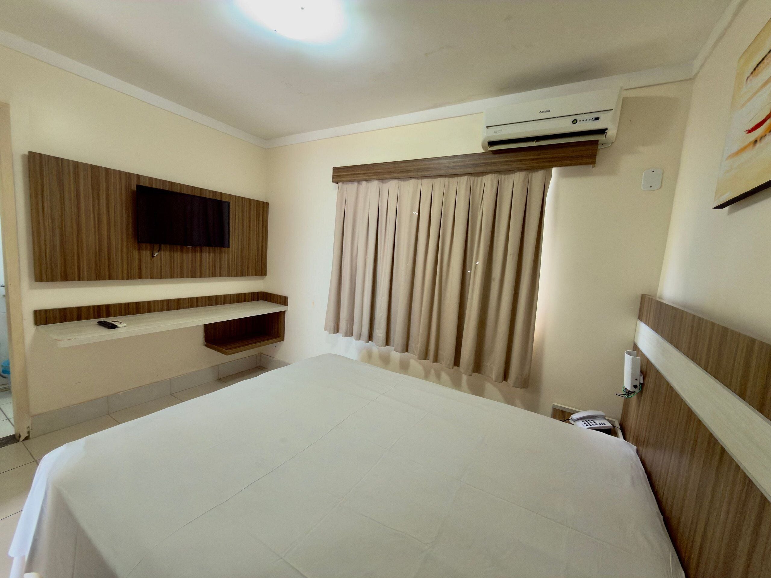 Quarto de hotel com cama, televisão montada, prateleira embutida, aparelho de ar condicionado e janela com cortinas.