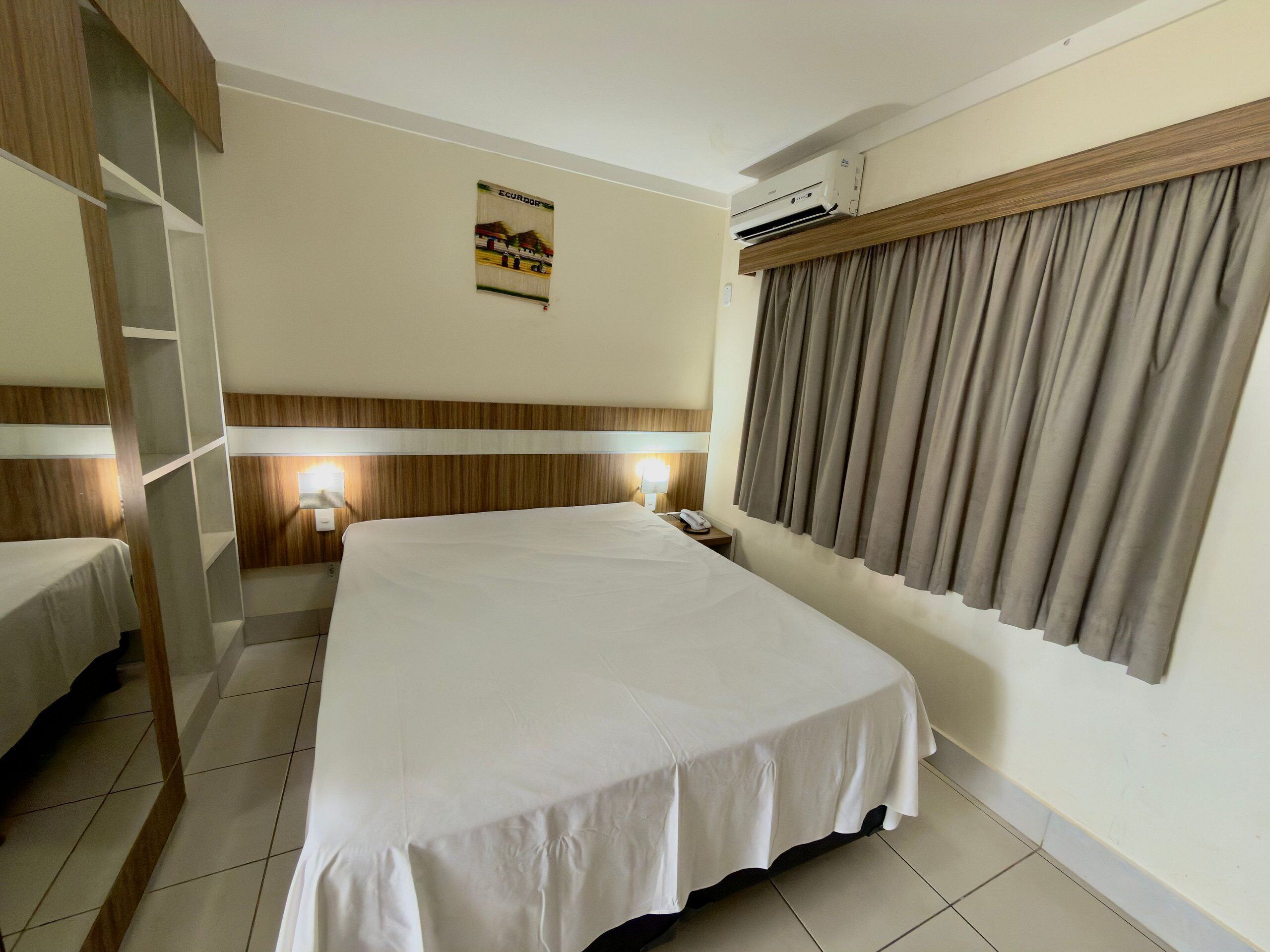 A hotel room with a large bed, a bedside table with a lamp, a wall-mounted air conditioner, and a poster on the wall.