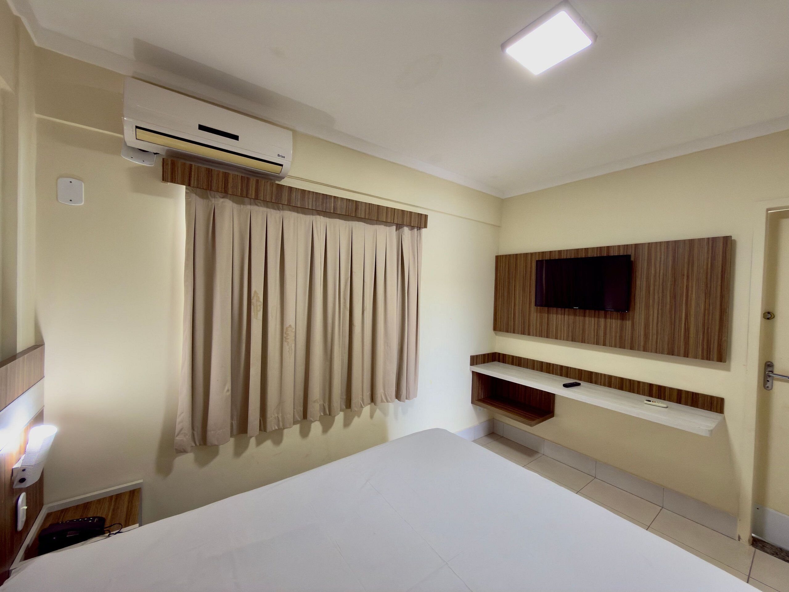 A hotel room with a bed, an air conditioning unit, a television mounted on the wall, and a curtain.