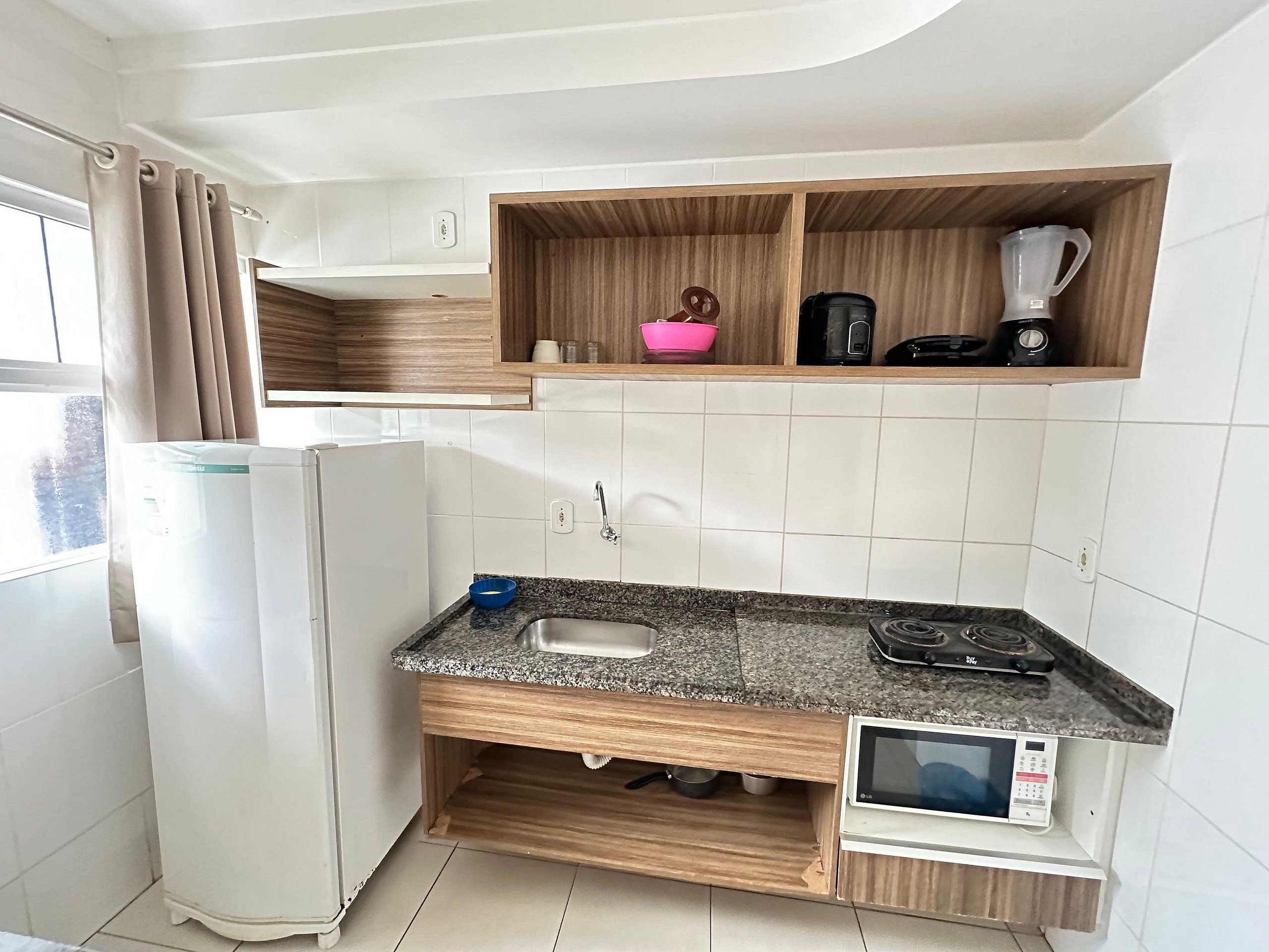 Une cuisine compacte équipée d’un réfrigérateur, d’un four à micro-ondes, d’un évier et d’étagères en bois au-dessus du plan de travail.