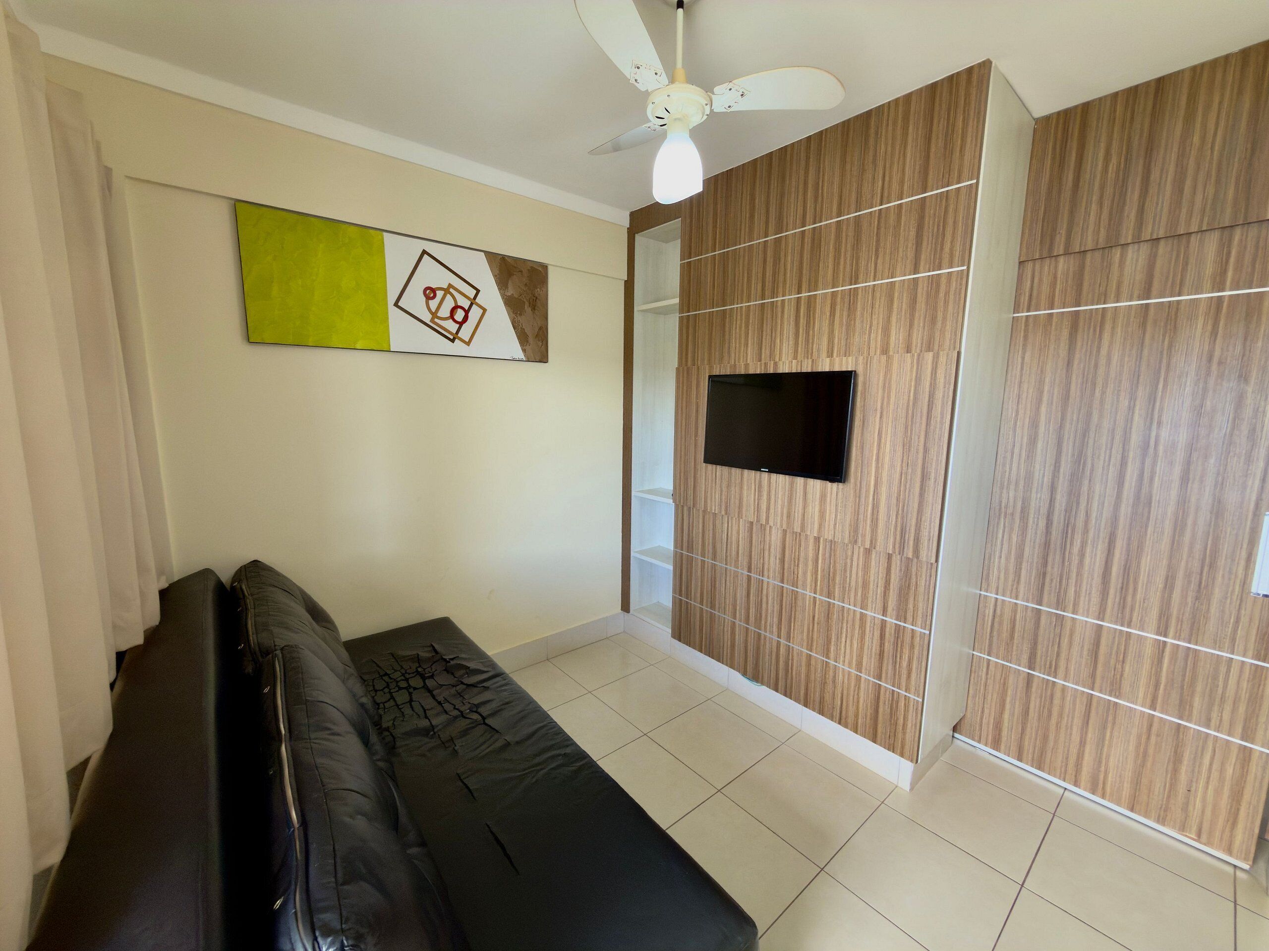 A living room with a black sofa, a flat-screen TV mounted on a wooden panel wall, a ceiling fan, and a large abstract painting.