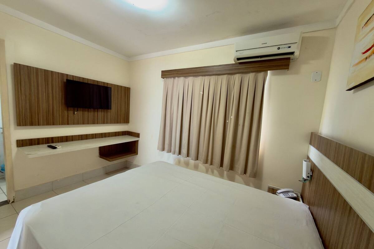Une chambre d’hôtel équipée d’un lit, d’un téléviseur fixé au mur, d’une étagère intégrée, d’un appareil de climatisation et d’une fenêtre avec des rideaux.