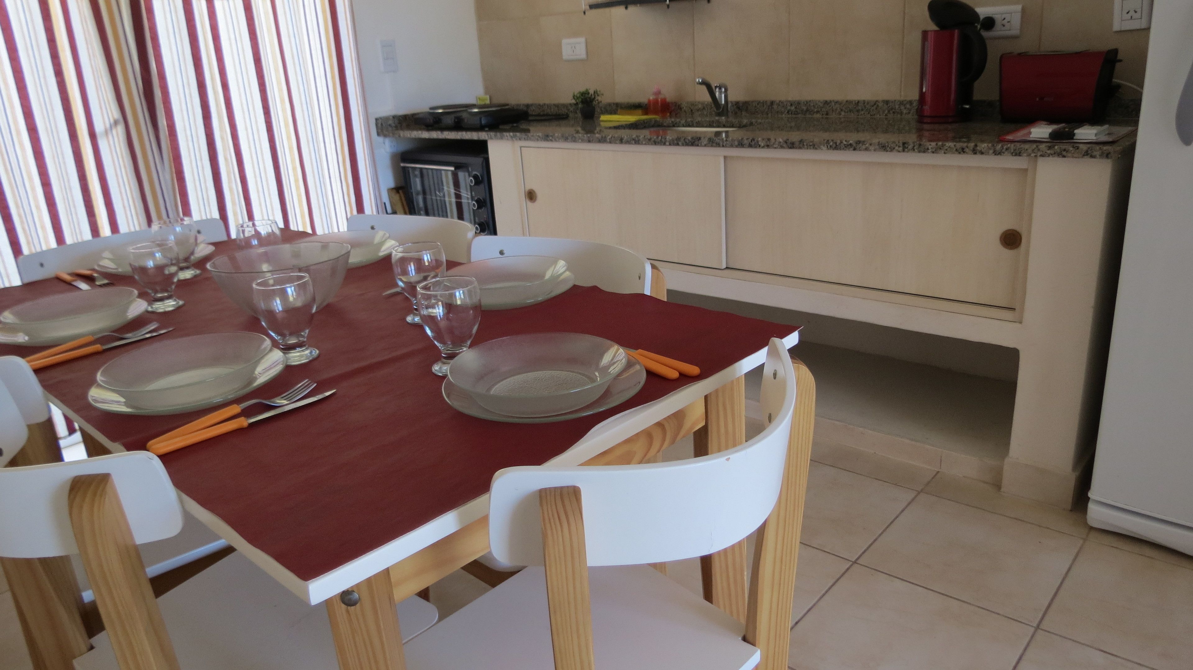A dining area with a red table set for six, white chairs, and a kitchen in the background.