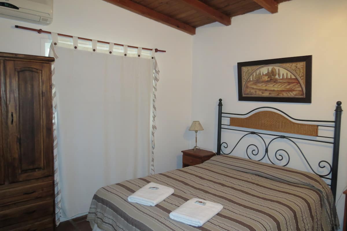 Un dormitorio con cama, armario de madera, mesita de noche con lámpara, un cuadro en la pared y una ventana con cortinas.