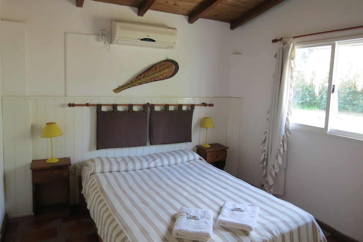 Un dormitorio con cama, dos mesitas de noche, aire acondicionado de pared, una pluma decorativa y una ventana con cortinas.