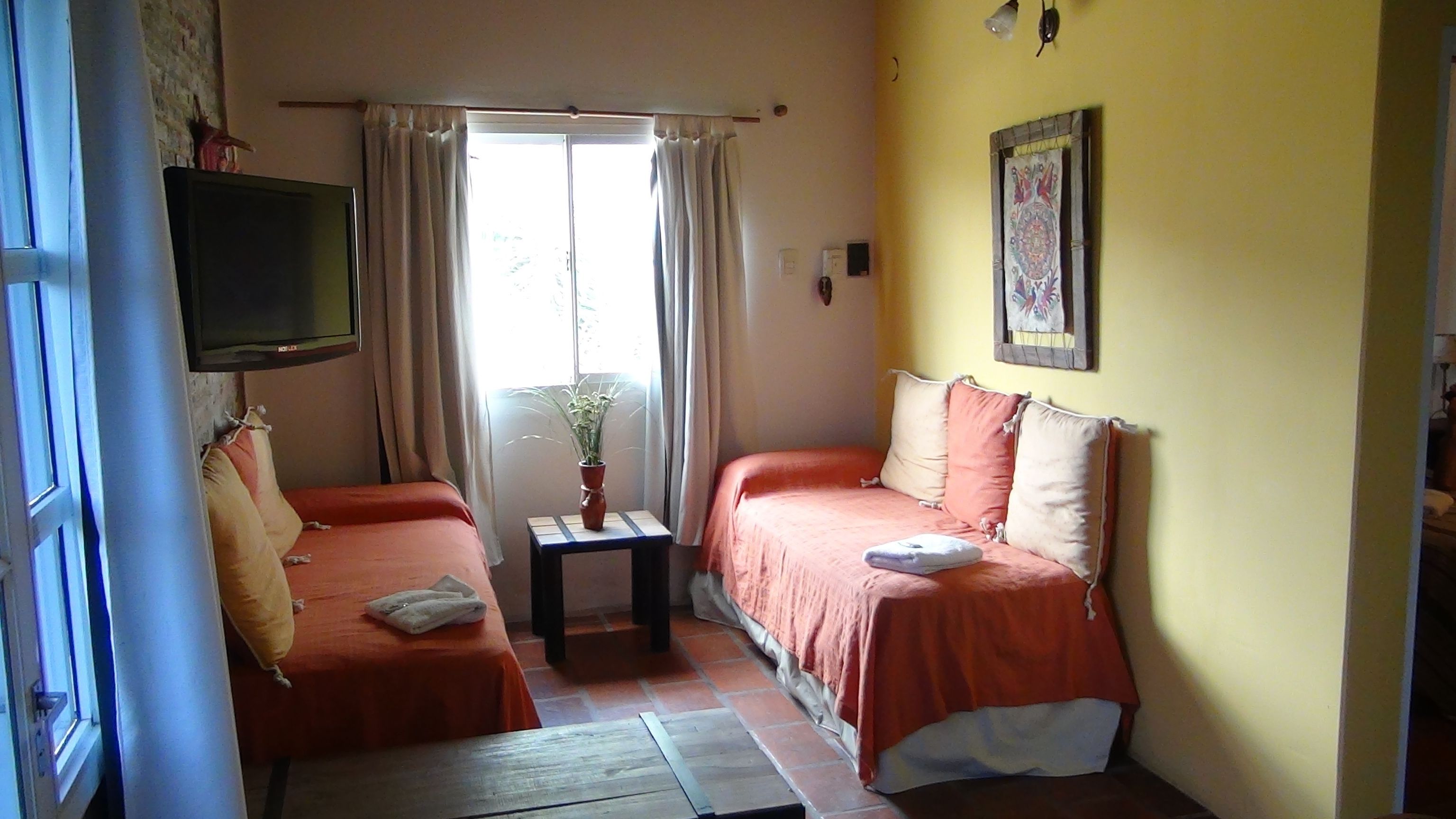 A room with two beds, a television mounted on the wall, a small table with a plant, and a window with curtains.
