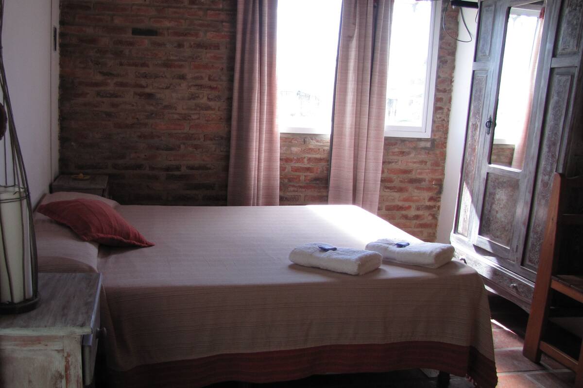 Un dormitorio con pared de ladrillo, una cama con almohada roja y una ventana con cortinas.