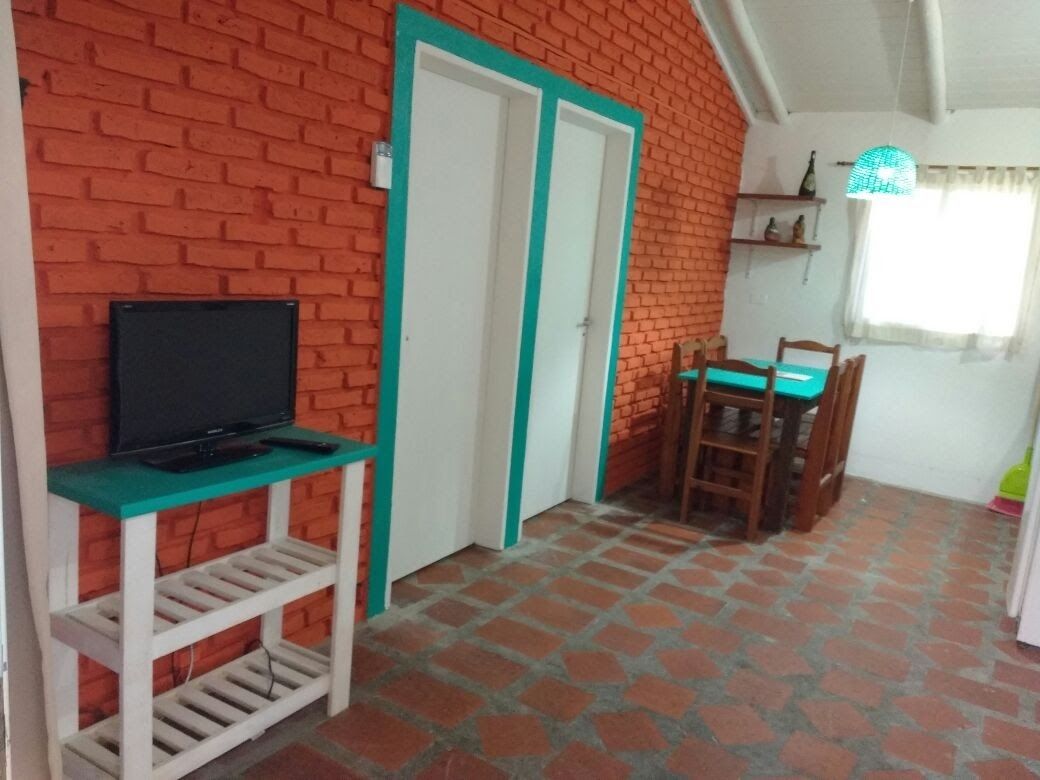 A room with a brick wall, a television on a white stand, a dining table with chairs, and a window with curtains.