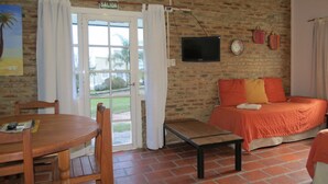 Una habitación con pared de ladrillo, cama con ropa de cama naranja, televisor montado en la pared y una puerta con la leyenda 'SALIDA'.
