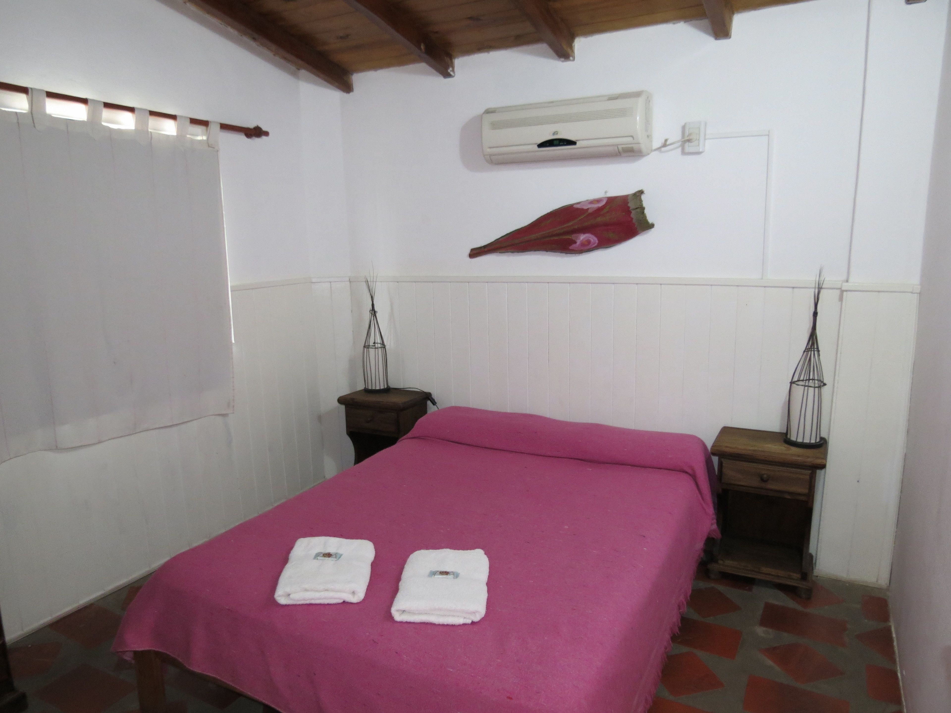 Una habitación con una cama, dos mesitas de noche, aire acondicionado de pared y una ventana con cortinas.