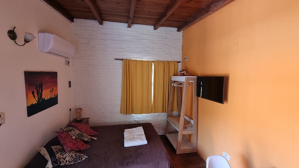 Quarto com cama, televisão montada na parede, janela com cortinas amarelas, ar condicionado fixado na parede e um quadro com paisagem desértica.
