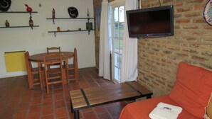 Un salón acogedor con pared de ladrillo, un televisor sobre un sofá, una mesa de madera con sillas y una ventana con cortinas.