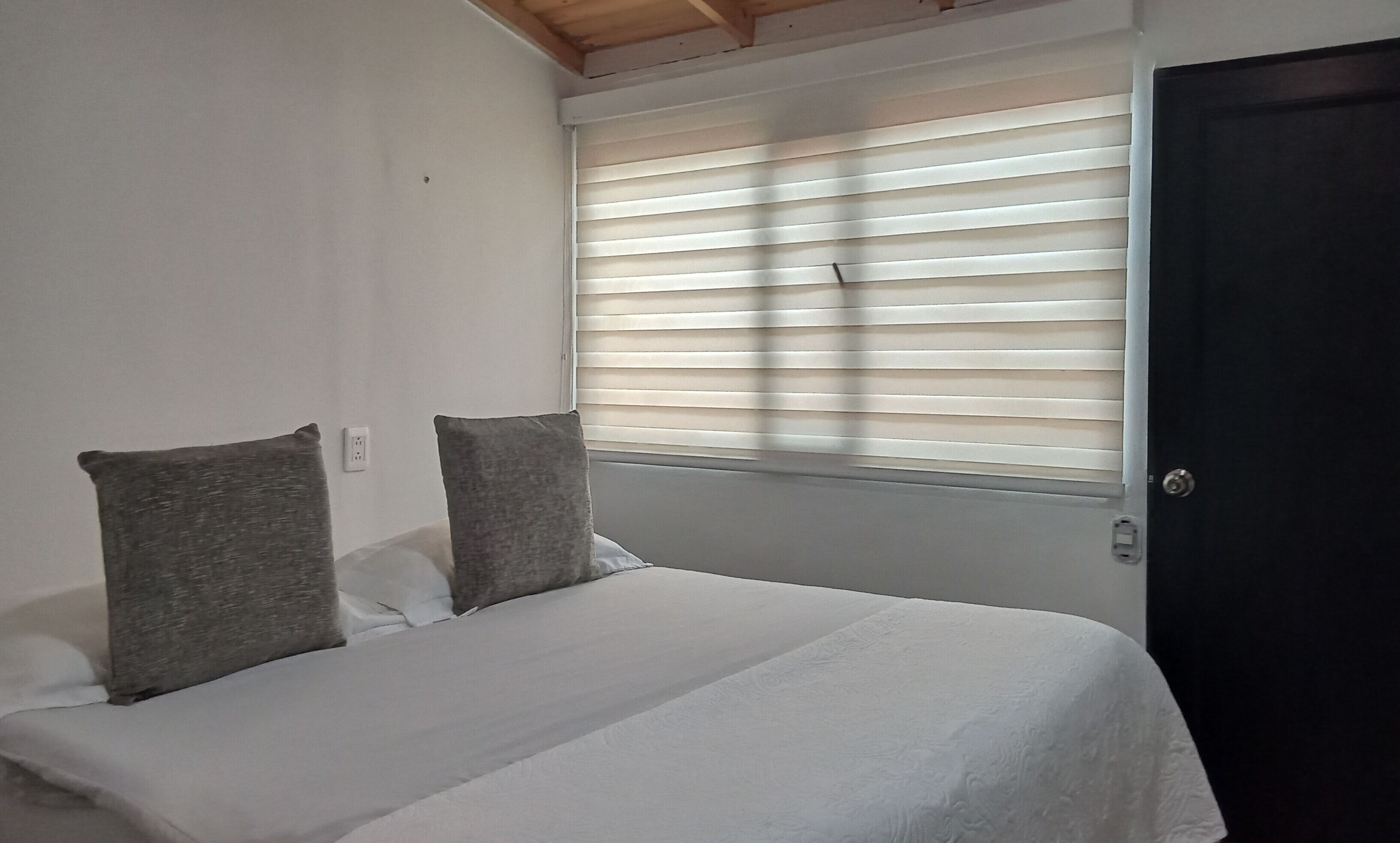 A bedroom with a bed, two pillows, a window with blinds, and a door.